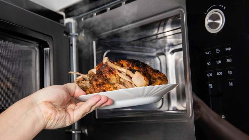 A hand removes a plate of rotisserie chicken from the microwave.