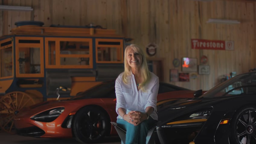 Woman sitting in chair with three McLarens parked in the background.