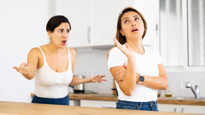 Frowning young Hispanic woman standing in home kitchen, listening listlessly to reprimanding from her displeased sister