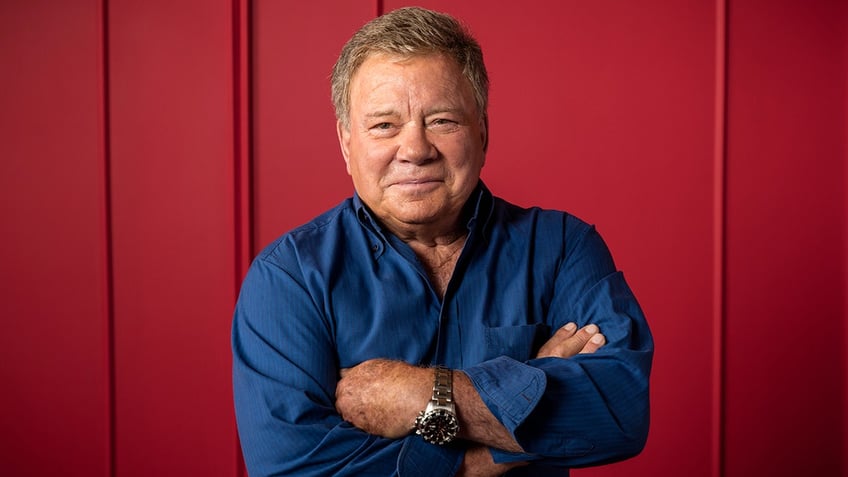 William Shatner in a blue shirt crosses his arms over his chest and soft smiles