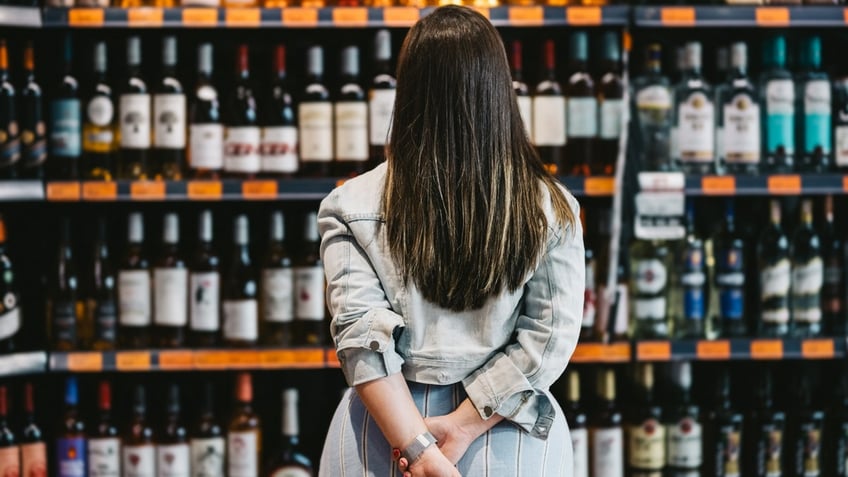 A woman stands with her hands behind her back as she faces a large selection of wines on shelves.