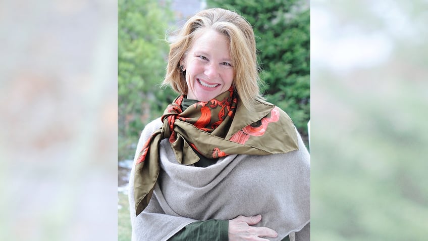 Lisa Brown Langley smiles while wearing a scarf and standing in a wooded area.
