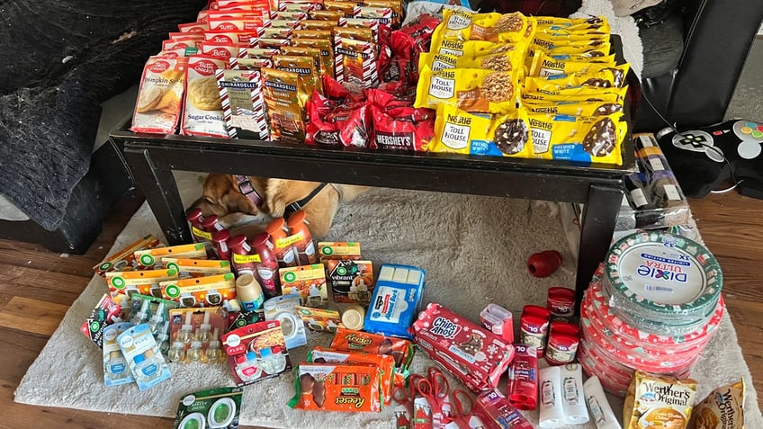 A dog sleeps under a table filled with grocery store purchases that were acquired by couponing. Items include chocolates, candies and cookie mixes.