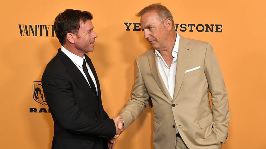 Taylor Sheridan in a black suit shakes the hand of Kevin Costner in a tan suit on the carpet at the "Yellowstone" premiere