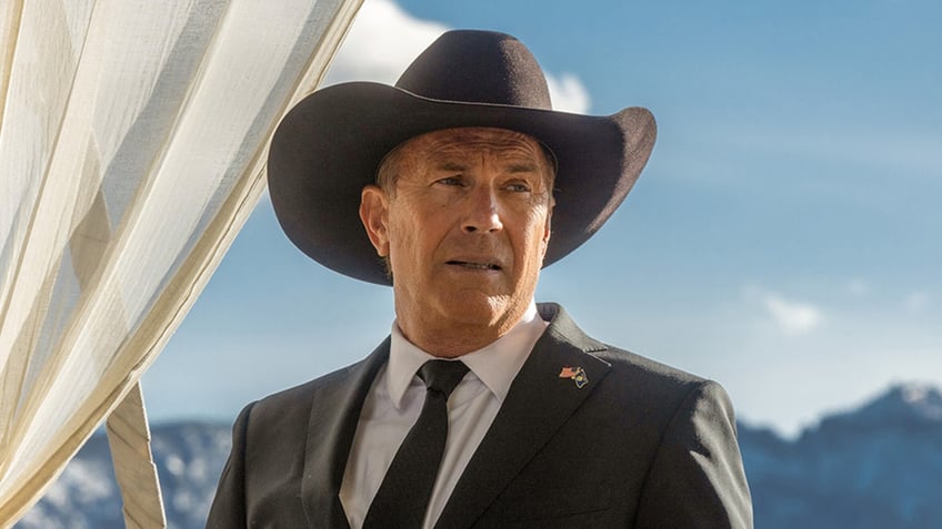 Kevin Costner in a black suit and black tie looks off in the distance wearing a cowboy hat for Yellowstone photo as John Dutton