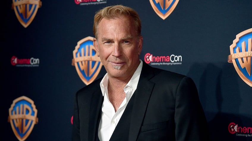 Kevin Costner in a white shirt slightly unbuttoned and black suit smirks on the carpet