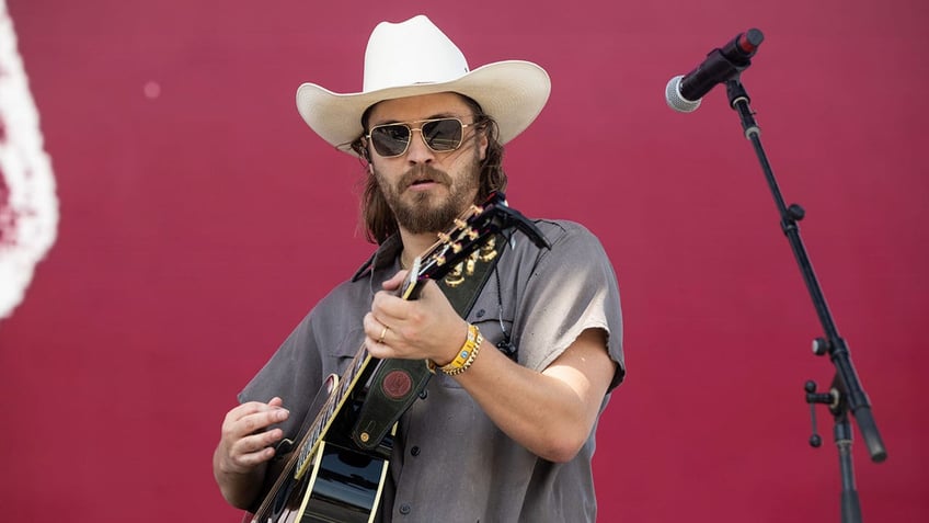 Luke Grimes in a grey shirt and off-white cowboy hat strums the guitar on stage