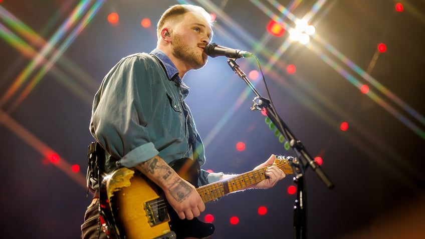 Zach Bryan wears a jean shirt and plays his guitar on stage